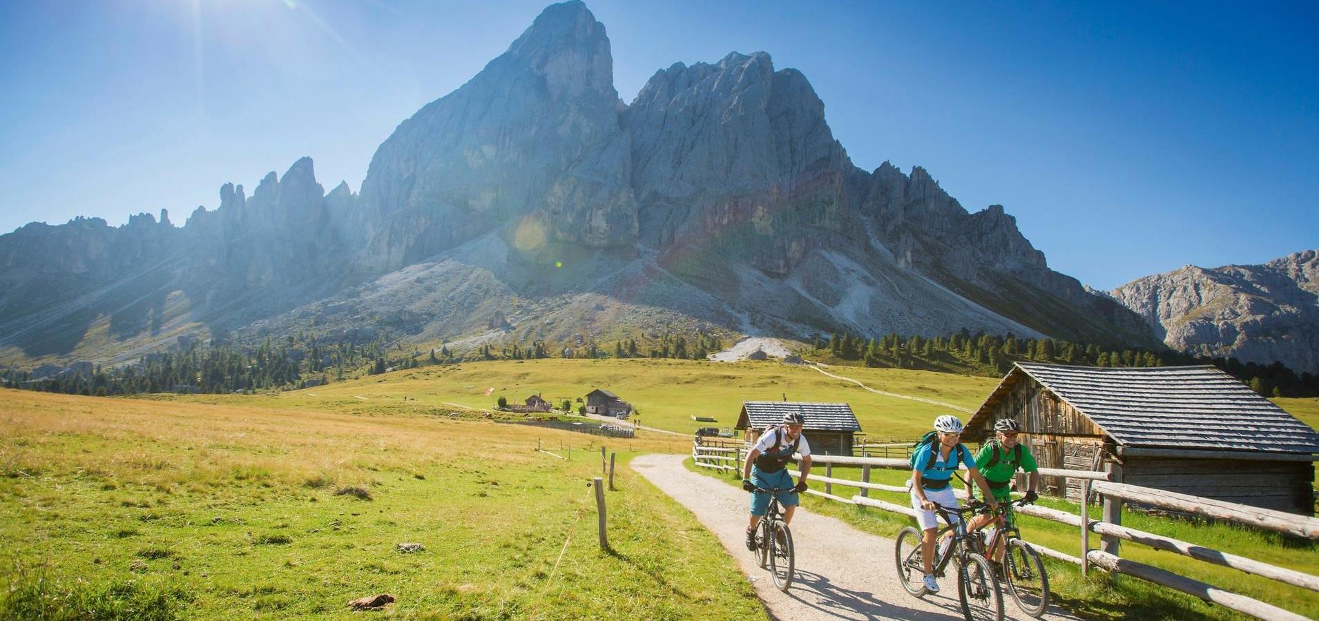 Tre ciclisti su un sentiero di montagna soleggiato, con baite e Dolomiti sullo sfondo.