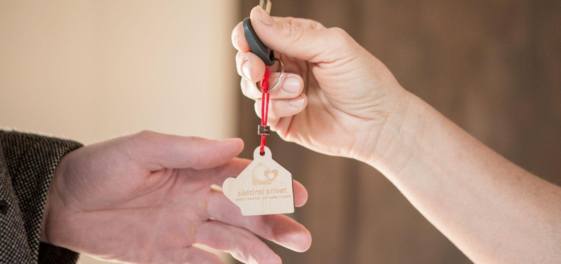Consegna delle chiavi con portachiavi in legno “südtirol privat” tra due mani in primo piano.