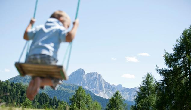In primo piano un bambino su un'altalena; sullo sfondo le Dolomiti.