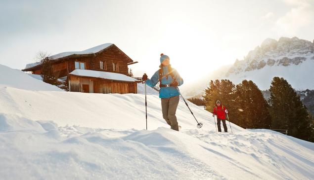 Due persone con racchette da neve davanti a una baita tra neve fresca e montagne innevate.