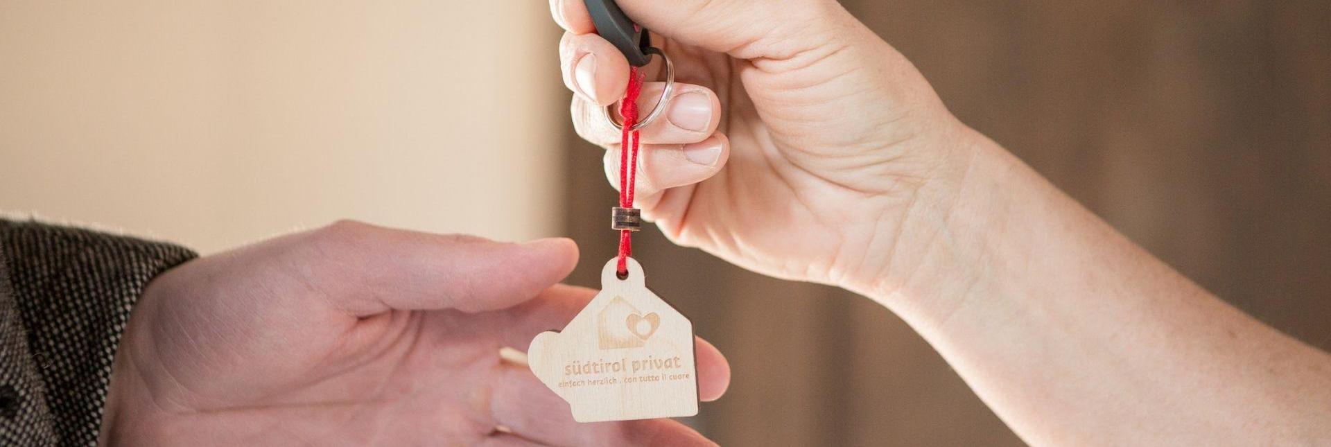 Consegna delle chiavi con portachiavi in legno “südtirol privat” tra due mani in primo piano.